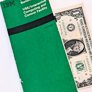 May include: A green IBM reference manual titled "VSE/Interactive Computing and Control Facility" with black text and a black stripe. A one-dollar bill is partially visible next to the manual.
