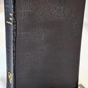 May include: A black hardcover book titled "The New Testament" with gold lettering and a gold emblem. The book has a textured cover and pink-colored edges. A one-dollar bill is visible in the foreground.