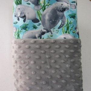 May include: A gray and blue fabric baby blanket with a pattern of manatees and fish. The blanket is made of soft, plush fabric and has a gray minky backing.