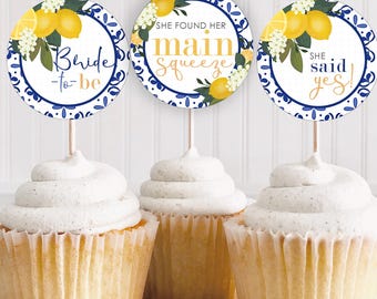 Puede incluir: Tres cupcakes con adornos de papel redondos que presentan limones amarillos, hojas verdes y patrones de azulejos azules y blancos. Los adornos muestran los textos Bride-to-be, She found her main squeeze y She said yes en azul y dorado.