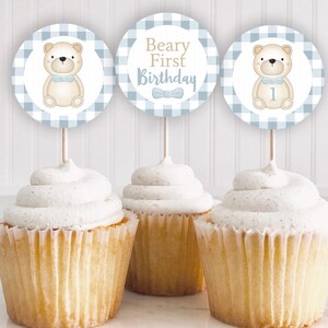 Puede incluir: Tres cupcakes con adornos. Los adornos son redondos con un patrón de cuadros azules y blancos. Dos adornos presentan una ilustración de oso de peluche, y uno dice "Beary First Birthday". Los cupcakes tienen glaseado blanco.