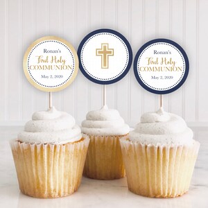 Pode incluir: Três cupcakes com cobertura branca, cobertos com toppers redondos de papel para cupcakes. Os toppers são azul marinho com letras douradas e uma cruz dourada. O texto nos toppers diz "Ronan's First Holy Communion May 2, 2020".