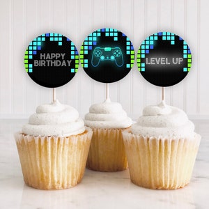 May include: Three cupcakes with black and green frosting toppers. The toppers have a pixelated design with a blue and green video game controller and the text "Happy Birthday", "Level Up".