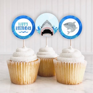 May include: Three cupcakes with blue and white shark themed cupcake toppers. The toppers say "Happy Birthday" and feature a shark with an open mouth and a shark with a closed mouth.