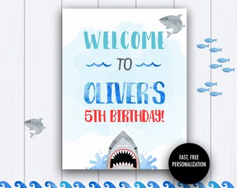 Op de afbeelding: Een blauw en wit welkomsbord met de tekst "Welkom bij Oliver's 5e verjaardag!" met een haai-illustratie. Het bord heeft een aquarel achtergrond en een rand van blauwe golven.