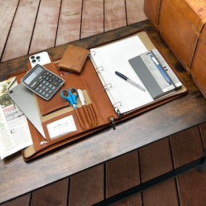 Könnte beinhalten: Ein braunes Lederportfolio mit Reißverschluss, das einen Taschenrechner, ein Smartphone, einen Notizblock, einen Stift, eine Schere und eine Visitenkarte enthält. Das Portfolio ist auf einem Holztisch geöffnet.