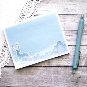 Puede incluir: Una tarjeta de acuarela azul con una silueta de ciervo blanco en una escena de bosque nevado. La tarjeta está en blanco y lista para un mensaje.
