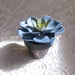 Puede incluir: Una planta suculenta artificial azul y verde en una maceta negra con un diseño decorativo rosa. La suculenta tiene múltiples capas de pétalos azules con centros verdes y amarillos. La maceta es negra con un patrón de remolino rosa.