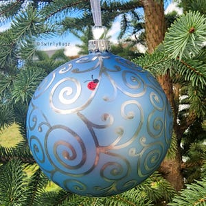 May include: A light blue Christmas ornament with silver swirl designs and a small red ladybug. The ornament is hanging from a silver-colored cap and a white ribbon. The text "SwirlyBugz" is visible on the ornament.
