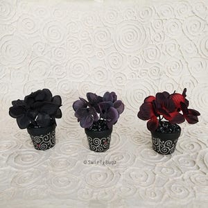 May include: Three small potted plants with artificial flowers in shades of black, purple, and red. The black pots have a decorative white swirl design and red accents. The background is a white textured surface.