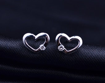 May include: A pair of silver heart-shaped stud earrings. Each earring features a heart outline with a small, round, clear gemstone set in the center. The earrings are displayed on a black, textured surface.