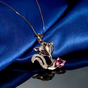 Puede incluir: Collar con colgante de zorro en oro rosa con una cadena fina. El zorro presenta un diseño brillante con piedras preciosas claras y rosas. El collar se muestra sobre una tela de satén azul.