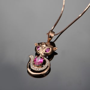 Puede incluir: Collar con colgante de gato en oro rosa con una piedra preciosa rosa y pequeñas gemas rosas para los ojos. La cola del gato se enrolla alrededor de una piedra preciosa rosa más grande, con una delicada cadena de oro rosa.