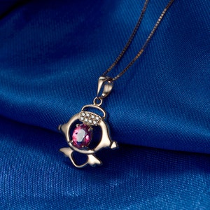 Puede incluir: Un collar con colgante dorado con una piedra preciosa rosa y pequeñas piedras blancas. El colgante tiene un diseño único y estilizado. La cadena del collar es delicada, sobre una tela azul oscuro.