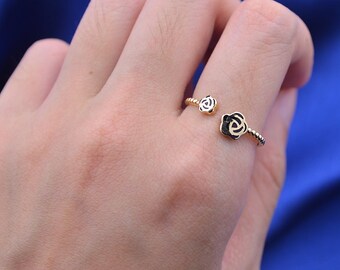 Può includere: Anello aperto color oro con due decorazioni a forma di rosa. Le rose hanno centri neri e petali bianchi, con un design a fascia intrecciata. L'anello è mostrato su un dito su sfondo blu.
