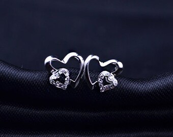 May include: A pair of silver-toned heart-shaped stud earrings. Each earring features an open heart design with a smaller heart-shaped cluster of clear gemstones at the center. The earrings are displayed on a dark, textured surface.