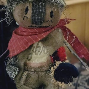May include: A fabric scarecrow doll with a brown hat, a red plaid scarf, and a burlap-textured body. The doll features stitched facial details, a patch on the hat, and a small sunflower accent. The chest area displays the cursive text "joy".