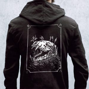 May include: Black hoodie with a white graphic print of a skull with teeth and plants on the back. The graphic is framed with a white border and geometric symbols.