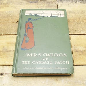 May include: A vintage hardcover book titled "Mrs. Wiggs of the Cabbage Patch" with a green cover. The cover features an illustration of a woman in a red dress and a landscape scene. The book is resting on a wooden surface.