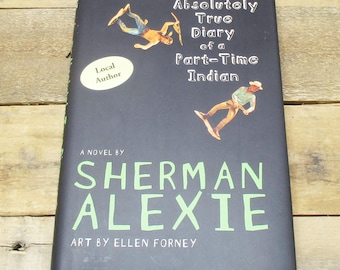 May include: A hardcover book titled "The Absolutely True Diary of a Part-Time Indian" by Sherman Alexie. The cover is black with illustrations and text in white and green. The book is on a wooden surface.