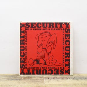 May include: A red square with black text and a cartoon illustration. The text reads "SECURITY IS A THUMB AND A BLANKET" around the border. The illustration depicts a character holding a blanket.