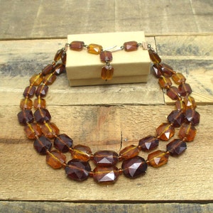May include: A three-strand amber glass necklace with faceted beads. The necklace is displayed on a wooden surface with a small box.