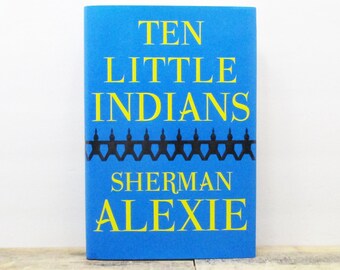May include: A hardcover book titled "Ten Little Indians" by Sherman Alexie. The book has a bright blue cover with yellow text and a black silhouette design. The book is displayed on a wooden surface.