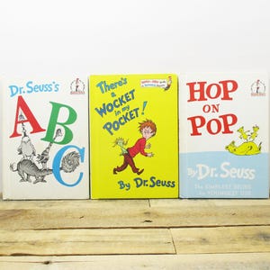May include: Three Dr. Seuss books are displayed on a wooden surface. The books are titled "Dr. Seuss's ABC", "There's a Wocket in my Pocket!", and "Hop on Pop". The covers feature colorful illustrations and text.