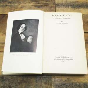 May include: An open book with a black and white portrait of a man on the left page and text on the right page. The text reads "DICKENS: A PORTRAIT IN PENCIL" by Ralph Straus. The book is on a wooden surface.