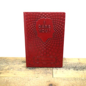 May include: A red journal with the words "I DARE YOU!" embossed on the cover. The cover has a geometric pattern and a silhouette of a face. The journal is on a wooden surface.