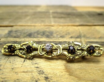 May include: Gold-tone brooch with a delicate design featuring three black gemstones and a clear rhinestone in the center.