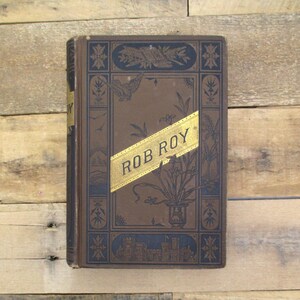 May include: Antique hardcover book titled "Rob Roy" with a brown cover and gold lettering. The cover features ornate blue and gold decorative elements, including floral and architectural designs. The book is displayed on a wooden surface.