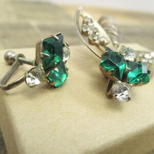 May include: A pair of vintage clip-on earrings with green and clear rhinestones. The earrings have a silver tone metal base.