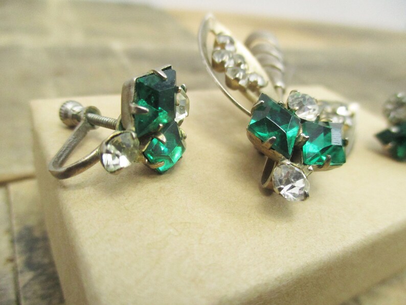 May include: A pair of vintage clip-on earrings with green and clear rhinestones. The earrings have a silver tone metal base.