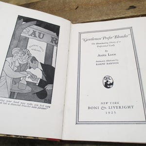 May include: Open book with black and white illustrations and text. The left page features a cartoon of people at a bar with the text "Kissing your hand may make you feel very good but a diamond bracelet lasts forever." The right page reads "Gentlemen Prefer Blondes" by Anita Loos.