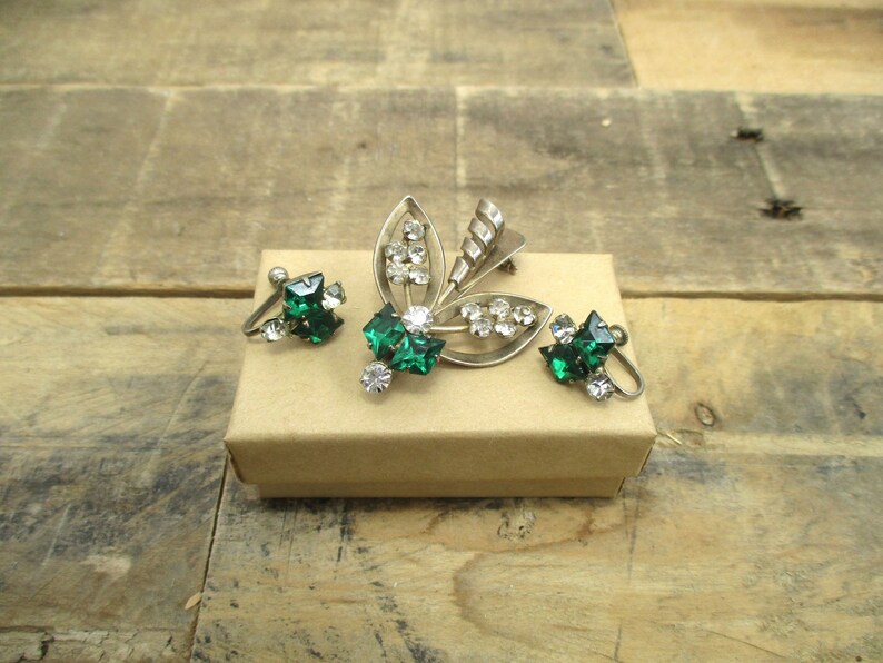 May include: A silver brooch and a pair of clip-on earrings with green and clear rhinestones. The brooch is shaped like a stylized leaf with a swirl design. The earrings are square shaped.