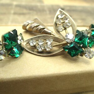 May include: A pair of vintage earrings with a silver tone metal base and green and clear rhinestones. The earrings feature a leaf design with a cluster of green rhinestones and a smaller cluster of clear rhinestones.