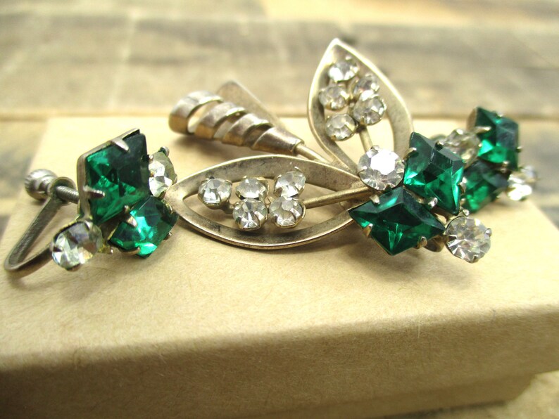 May include: A pair of vintage earrings with a silver tone metal base and green and clear rhinestones. The earrings feature a leaf design with a cluster of green rhinestones and a smaller cluster of clear rhinestones.