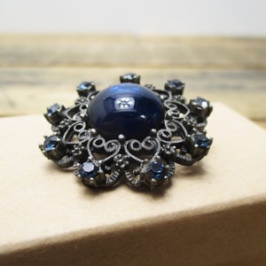 May include: A vintage brooch with a large, dark blue cabochon stone surrounded by a filigree design and smaller blue rhinestones.