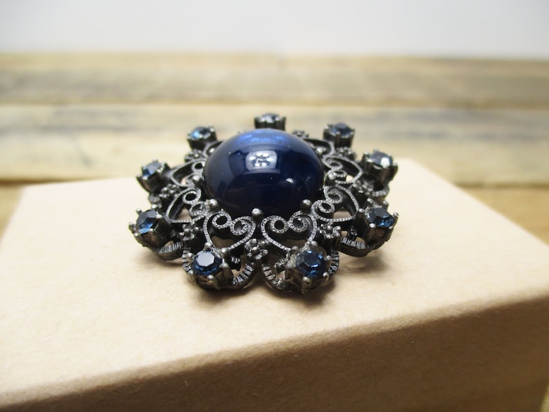 May include: A vintage brooch with a large, dark blue cabochon stone surrounded by a filigree design and smaller blue rhinestones.