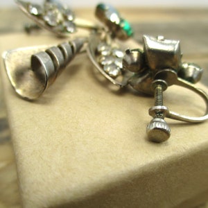 May include: Silver tone earrings with a spiral design and clear and green rhinestones. The earrings have a screw-back closure.