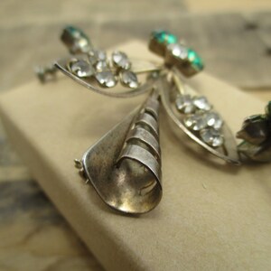 May include: A silver brooch with a three-dimensional design featuring a central cone shape with a swirl pattern and two wings with clear and green rhinestones.