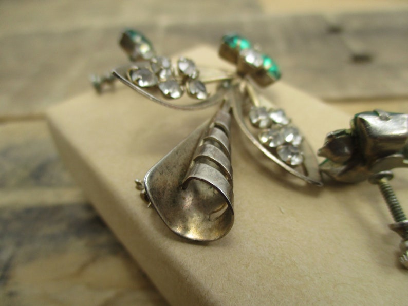 May include: A silver brooch with a three-dimensional design featuring a central cone shape with a swirl pattern and two wings with clear and green rhinestones.