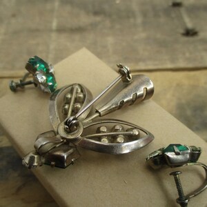 May include: A silver tone brooch and matching earrings with green glass stones. The brooch is shaped like a stylized flower with a pin on the back. The earrings are clip-on style.