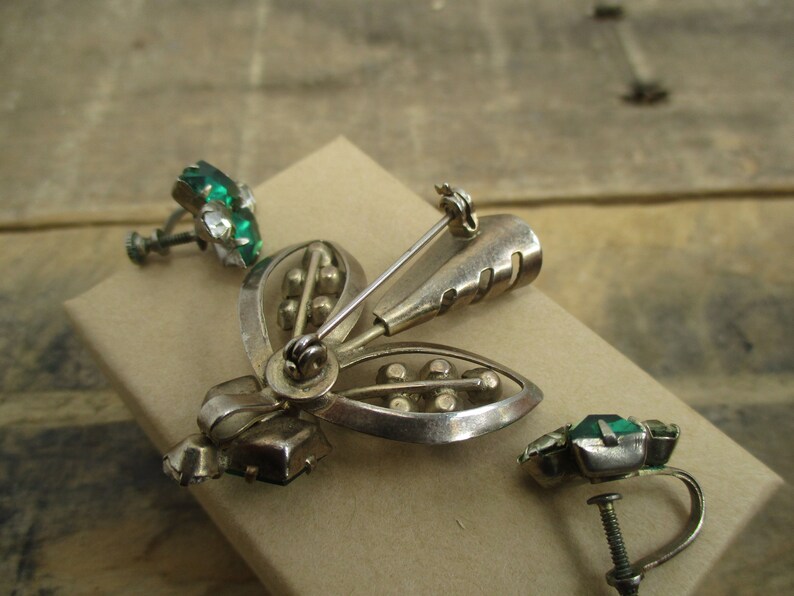 May include: A silver tone brooch and matching earrings with green glass stones. The brooch is shaped like a stylized flower with a pin on the back. The earrings are clip-on style.