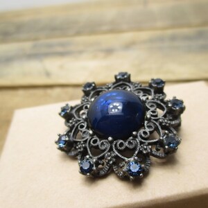 May include: A dark blue glass cabochon set in a silver filigree brooch with smaller blue rhinestones around the edge.