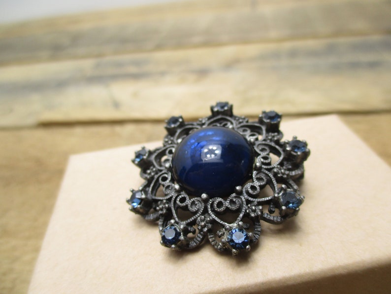 May include: A dark blue glass cabochon set in a silver filigree brooch with smaller blue rhinestones around the edge.