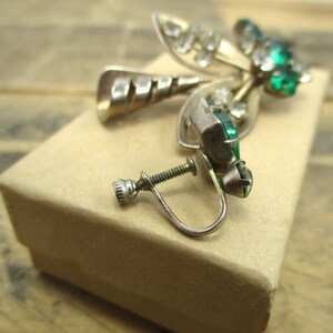 May include: A pair of silver tone clip-on earrings with green and clear rhinestones. The earrings are in the shape of a leaf with a swirl design.