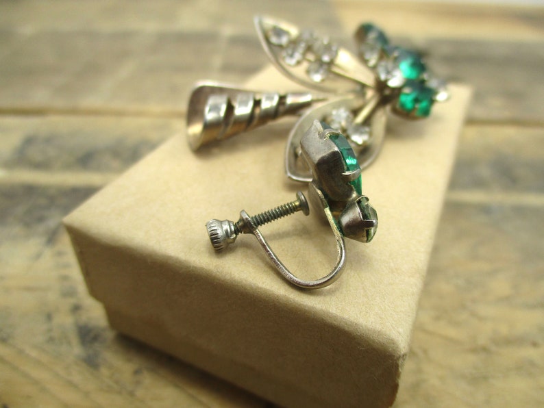 May include: A pair of silver tone clip-on earrings with green and clear rhinestones. The earrings are in the shape of a leaf with a swirl design.