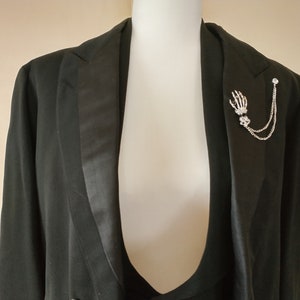 May include: A black tuxedo jacket with a silver skeleton hand lapel pin. The pin has a chain attached to it.